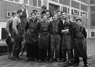 Das Bild zeigt eine Gruppe junger Männer und Frauen in Arbeitskleidung, die vor einem Gebäude posieren. Sie wirken freundlich und stolz.
