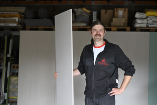 Ein Handwerker steht in einer Werkstatt und hält ein großes Plattenteil in der Hand. Im Hintergrund sind Materialien und Werkzeuge sichtbar.