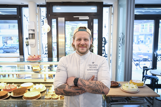 Ein lächelnder Koch steht mit verschränkten Armen vor einer Vitrine voller leckerer Kuchen und Torten in einem Café.