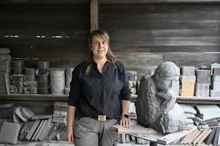 Eine Künstlerin steht in ihrer Werkstatt, neben einer Steinfigur. Im Hintergrund sind diverse Steinmaterialien sichtbar. Sie wirkt konzentriert und kreativ.
