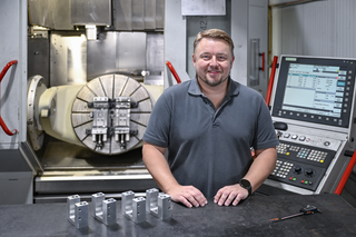 Der Mann steht an einem Maschinenarbeitsplatz mit einer modernen CNC-Fräse. Vor ihm liegen mehrere metallische Bauteile.