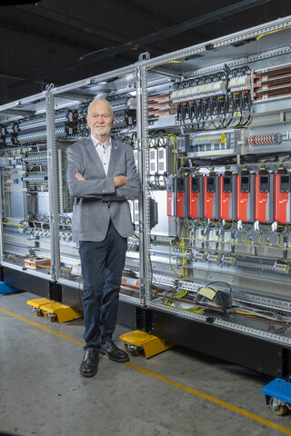 Ein lächelnder Mann steht vor einem großen, technisch ausgestatteten Schaltschrank. Die Umgebung wirkt modern und industriell.