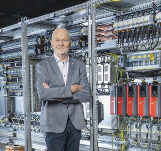 Ein lächelnder Mann steht vor einem großen, technisch ausgestatteten Schaltschrank. Die Umgebung wirkt modern und industriell.