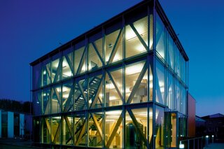 Das Bild zeigt ein modernes, transparenter Gebäude mit einer auffälligen Glasfassade und einem klaren, geometrischen Design bei Nacht.