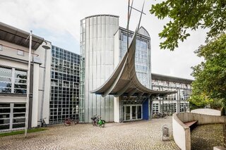 Das Bild zeigt ein modernes Gebäude mit Glasfassade und auffälligem Dach. Vor dem Eingang stehen Fahrräder, umgeben von Grünflächen.