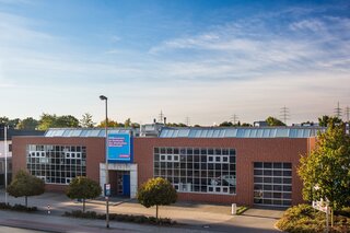Das Bild zeigt ein modernes, zweigeschossiges Gebäude mit großen Fenstern und einer rot gemauerten Fassade, umgeben von Bäumen und einem klaren Himmel.