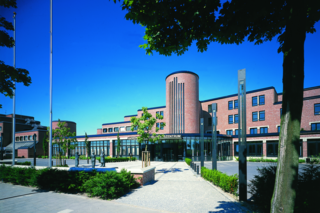 Das Bild zeigt ein modernes Gebäude mit einer markanten Fassade, umgeben von Bäumen und einem klaren blauen Himmel. Einladend und architektonisch ansprechend.