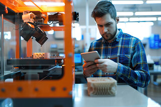 Ein junger Mann in einem karierten Hemd steht vor einem 3D-Drucker und schaut auf sein Handy, während er einen gedruckten Gegenstand überprüft.